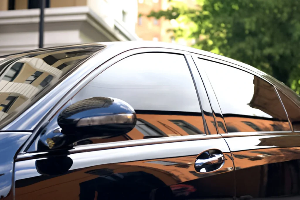 Glossy black car with tinted windows reflecting surrounding buildings and greenery, showcasing a sleek, polished appearance.