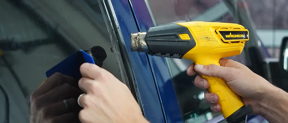 Close-up of hands applying window tint film to a car window using a yellow Wagner heat gun and a blue squeegee to smooth the material.