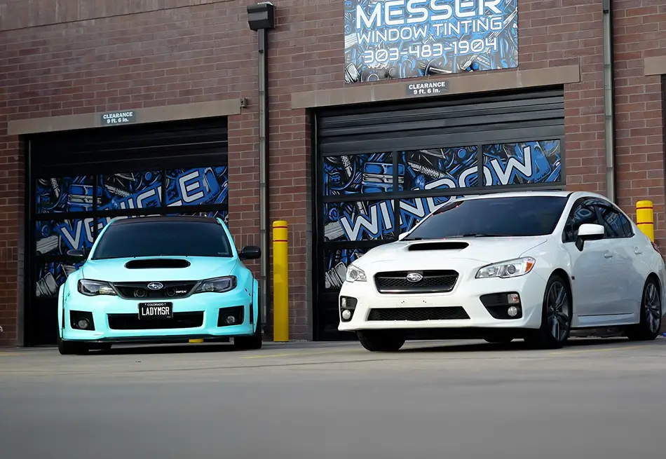 Two Subaru WRX sedans, one wrapped in teal and the other in white, parked outside Messer Window Tinting with tinted windows and custom vinyl graphics on garage doors.