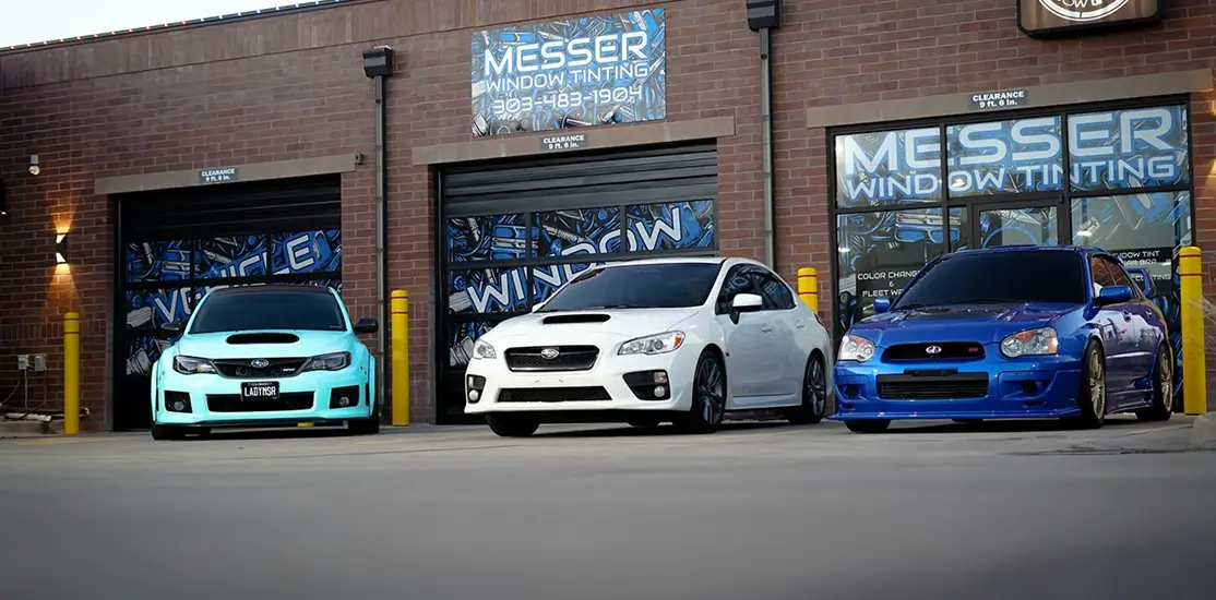 Three Subaru cars—mint green, white, and metallic blue—parked in front of Messer Window Tinting, a brick building with blue signage and custom window designs. The shop's name, phone number, and services like color changes and fleet work are displayed on the building.