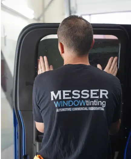 Technician wearing a “Messer Window Tinting” shirt applies tint film to the window of a vehicle, pressing it into place with both hands.