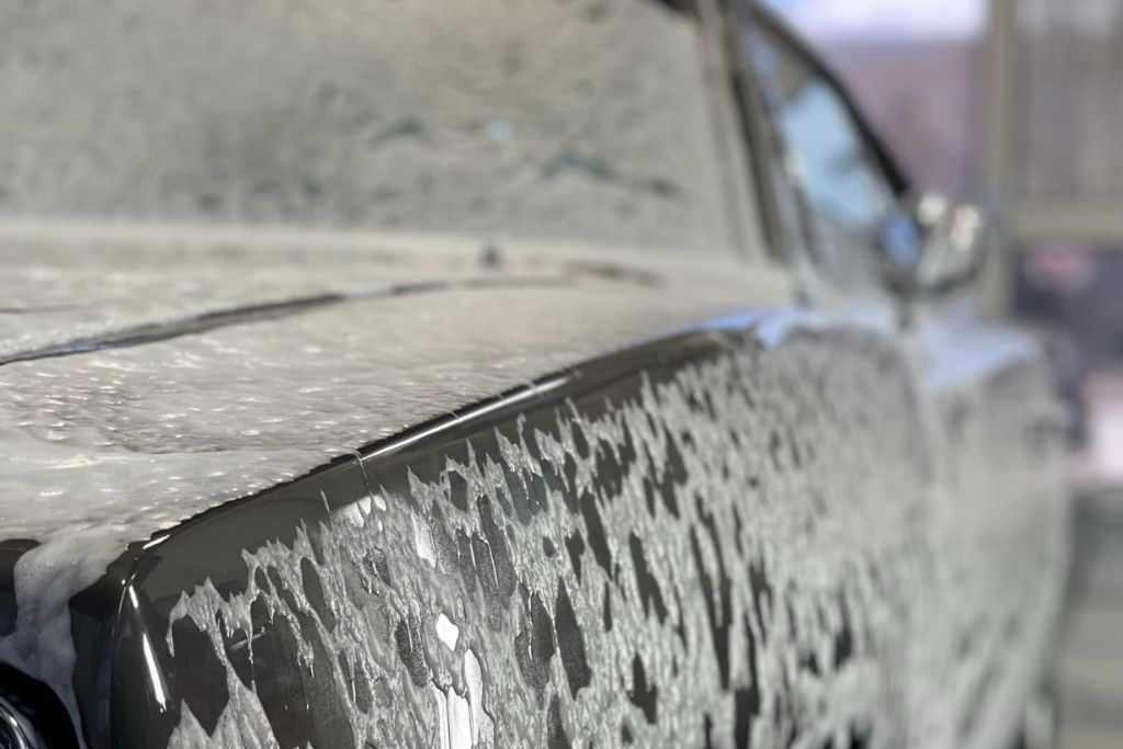 Close-up of a dark-colored car covered in thick, white foam during a wash, highlighting the importance of regular, gentle cleaning to maintain ceramic coating performance and hydrophobic effect.
