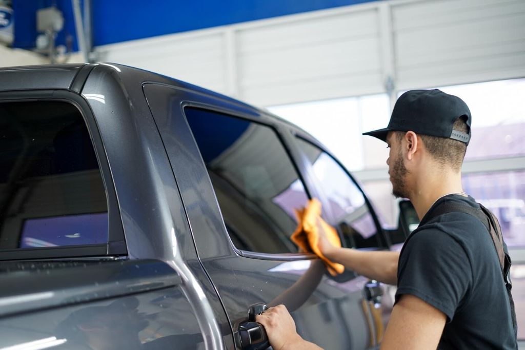 A man in a black shirt and cap wipes down the side of a clean gray truck with an orange microfiber cloth inside a well-lit garage, demonstrating the easy maintenance required for ceramic-coated vehicles.