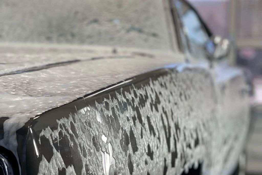 Close-up of a dark-colored car covered in thick white foam during a hand wash, highlighting the paint protection film's surface during cleaning. The image emphasizes the gentle washing method used to preserve the film's condition.