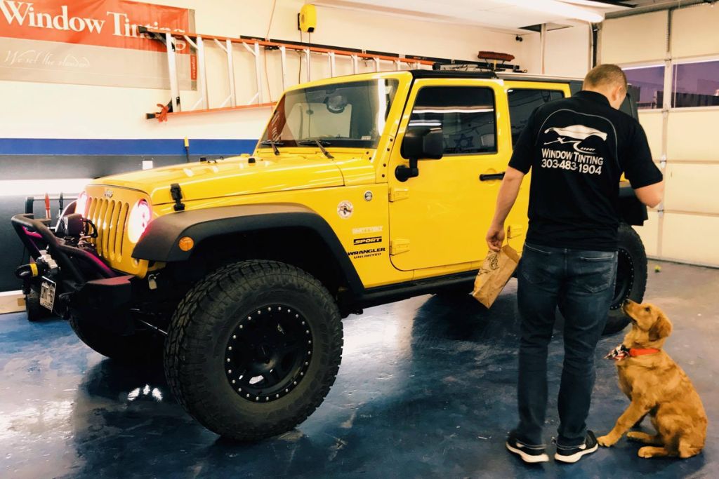 A yellow Jeep Wrangler is parked inside a window tinting shop, with a man in a "Window Tinting" shirt holding a paper bag while a golden retriever puppy sits attentively by his side.
