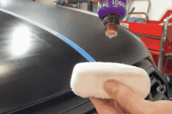 A person squeezes drops of ceramic coating liquid from a purple bottle onto a white applicator pad, preparing to apply it to a black car hood with a blue tape line down the center.