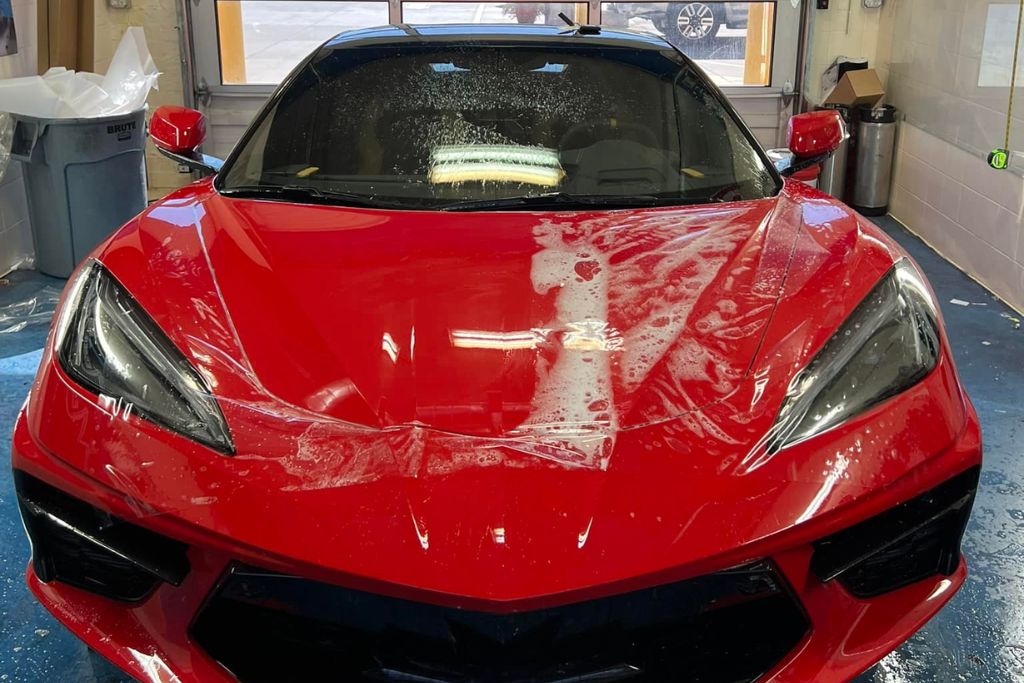 A red sports car in a detailing garage with paint protection film being installed on the hood, visible with water and air bubbles trapped underneath as part of the professional application process. The image highlights the precision required for expert PPF installation in Douglas County, USA.