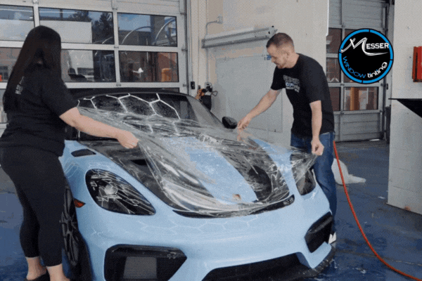 Two certified technicians at Messer Window Tinting carefully apply a large sheet of paint protection film to the hood of a light blue sports car inside a garage. The transparent film is being aligned before smoothing, showcasing professional PPF installation in Douglas County, USA.
