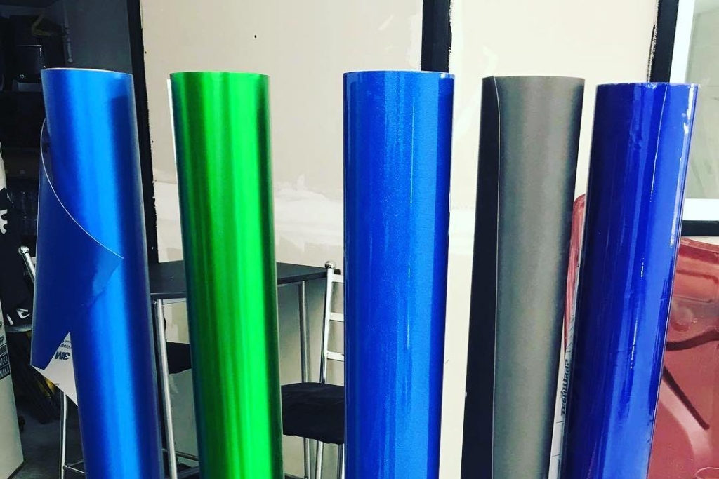 Five upright rolls of glossy and matte vinyl wrap in blue, green, and gray finishes displayed in a workshop, illustrating color and finish options when exploring what is vinyl wrapping for seasonal vehicle styling.
