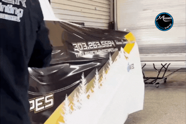 Technician from Messer Window Tinting applies a custom vinyl wrap with branding and mountain tree graphics to the side of a vehicle inside a workshop, showcasing professional wrap services in Parker.