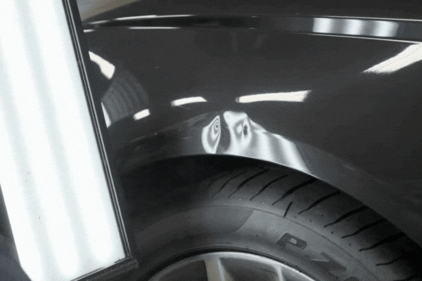 A close-up view of a car fender undergoing paintless dent repair, showing a technician using reflection board lighting to locate and fix a dent above the wheel well without damaging the original paint.