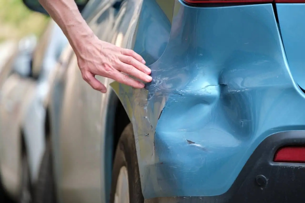 A close-up of a heavily dented and scratched rear quarter panel on a blue car, with a person's hand inspecting the damage that includes paint transfer, deep creases, and surface cracks—conditions unsuitable for paintless dent repair.