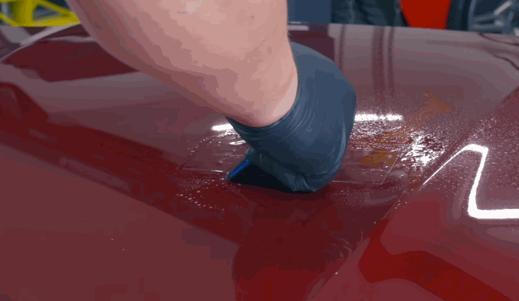 A gloved hand preparing a red car surface by using a clay bar or prep tool before applying ceramic coating.