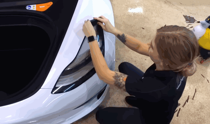 A woman kneels beside a white car and carefully applies black vinyl film to the headlight area using her fingers.