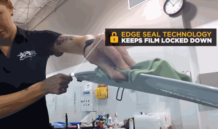 A technician stretches and presses a protective film along a car part while a graphic overlay reads “Edge Seal Technology Keeps Film Locked Down.”