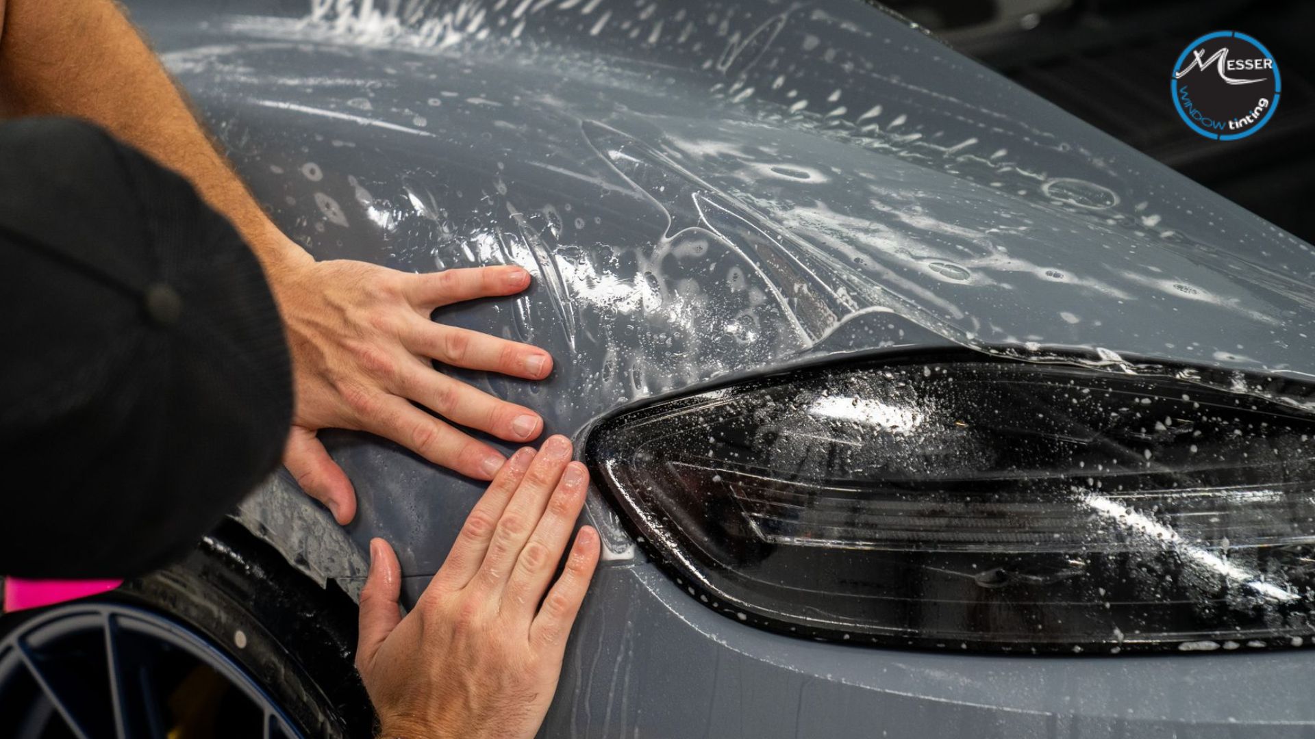 Close-up of a technician's hands applying clear paint protection film to the front fender and headlight of a gray car, smoothing it out with soapy water.