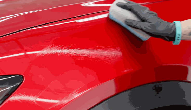 Hand in a black glove applying ceramic coating on a red car hood using a foam applicator pad, leaving visible streaks as the product is spread evenly.