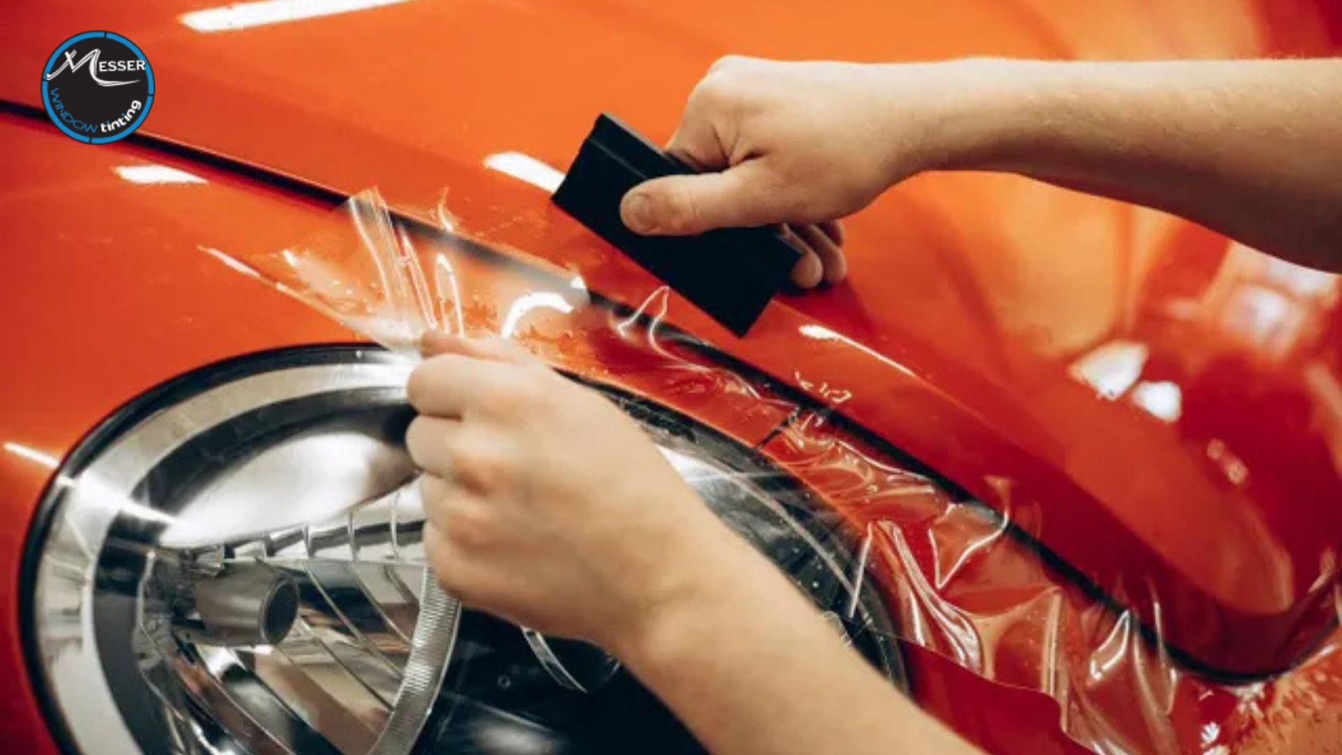 Hands applying clear paint protection film to the hood and headlight of a red car using a squeegee.