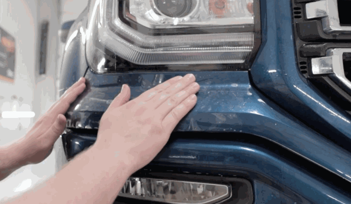 Hands pressing down a freshly applied paint protection film on the front bumper of a blue truck, smoothing it around the headlight and grille.