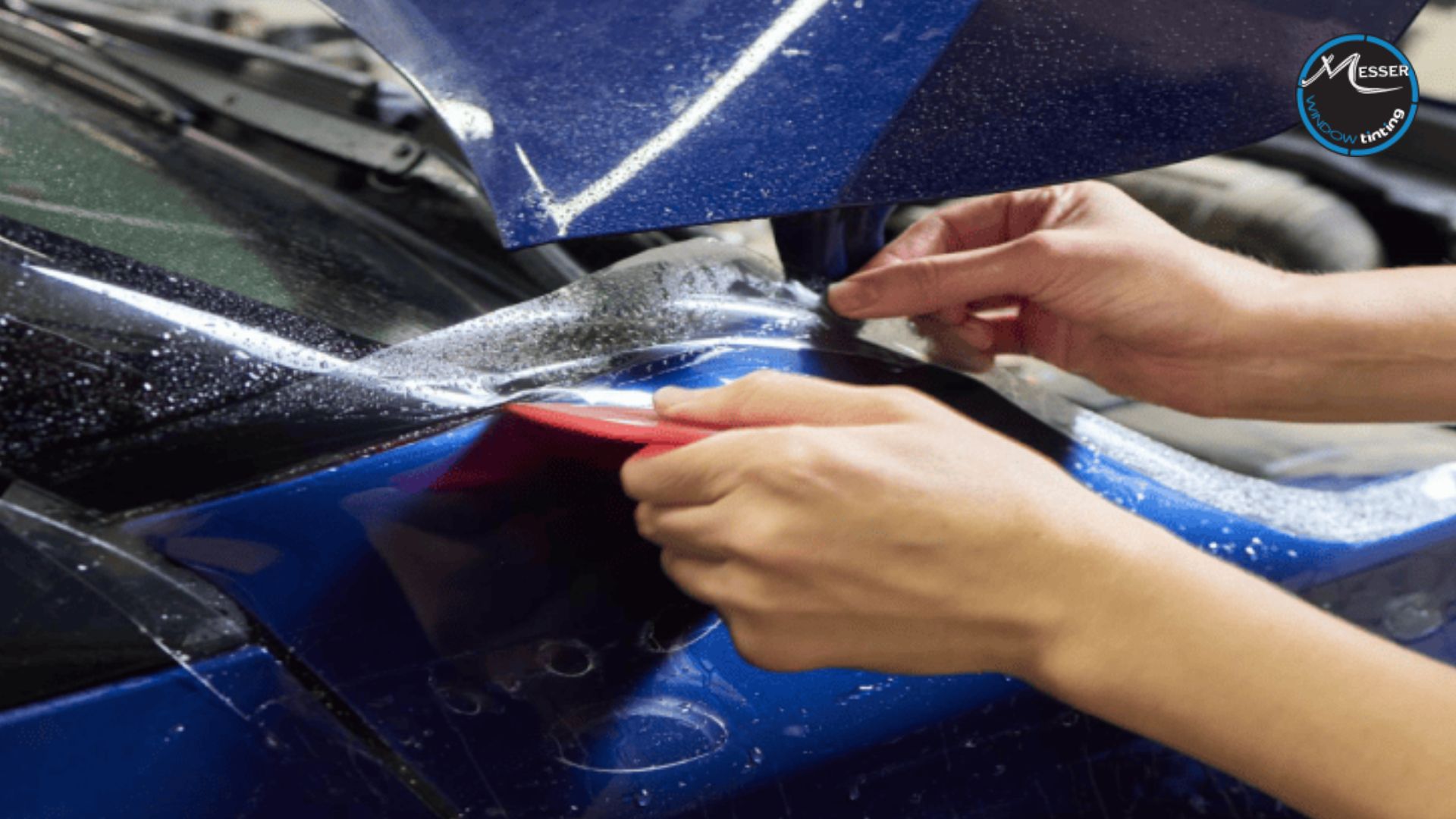 Close-up of hands applying paint protection film to the fender of a dark blue car using a red squeegee, with the film visibly wet for adjustment.