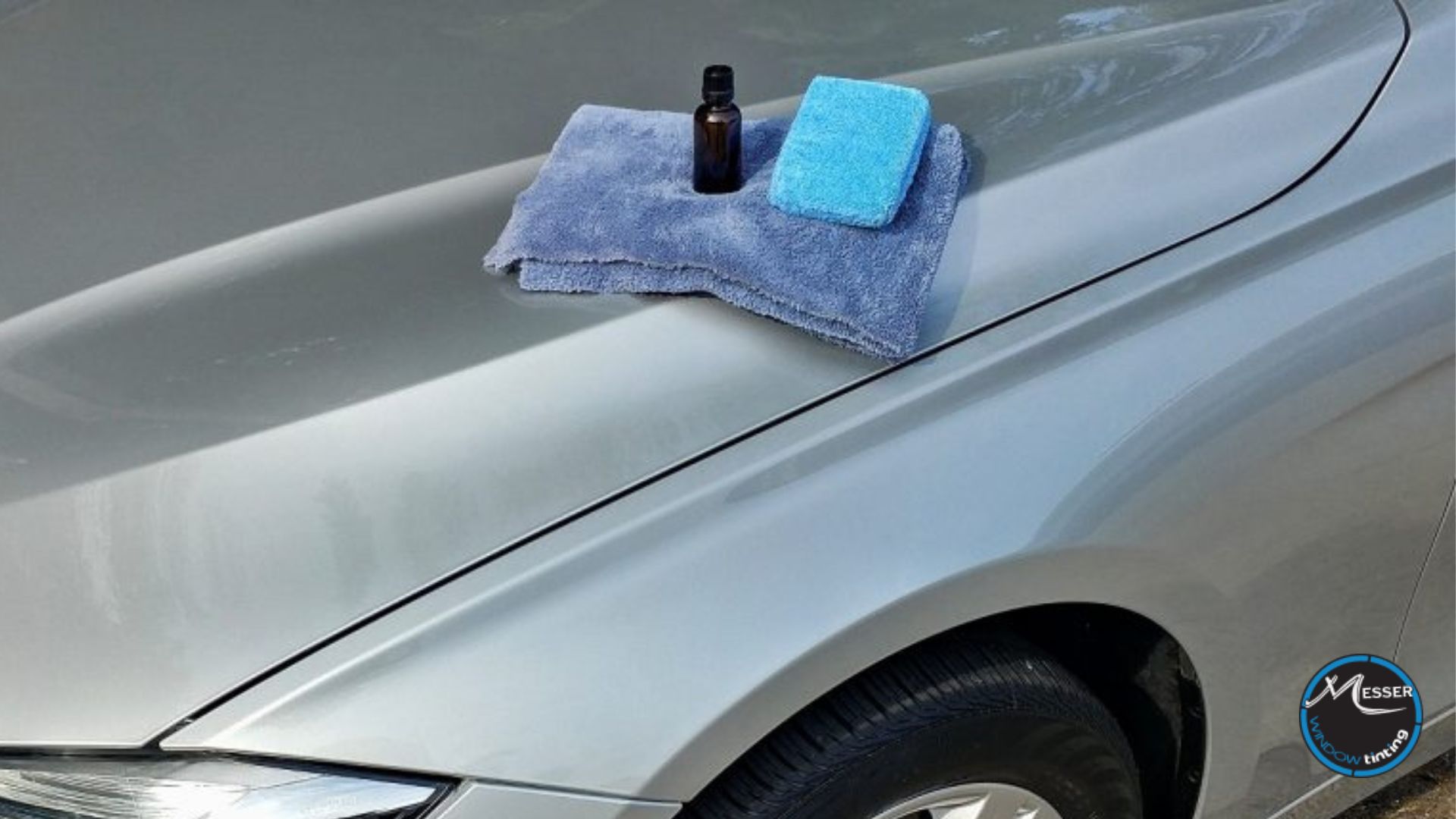 A bottle of ceramic coating solution, microfiber towels, and applicator pad placed on the hood of a silver car, ready for use.
