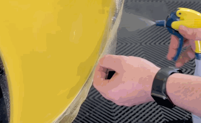 Close-up of paint protection film being carefully applied to the fender of a yellow car using a spray bottle and hand tension.