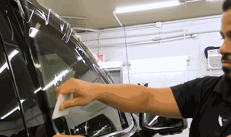 A detailer smooths freshly applied window tint film onto a car's side window using a white squeegee tool to remove bubbles.