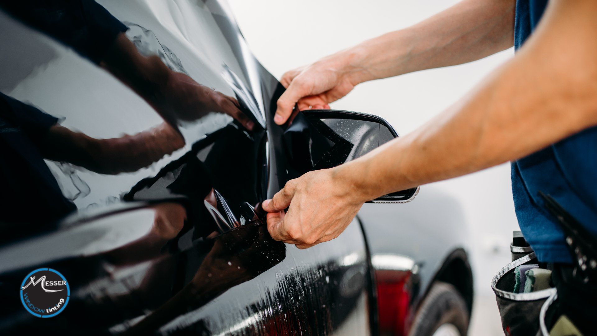 A close-up of a technician carefully installing dark window tint on a car's rearview mirror panel using a heat-shrunk film.
