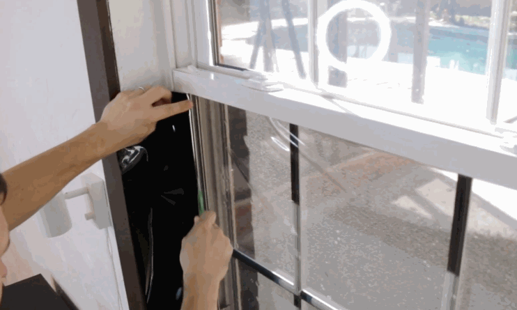 A person carefully aligns and presses window tint film onto a residential window, using a green squeegee tool to smooth the surface.