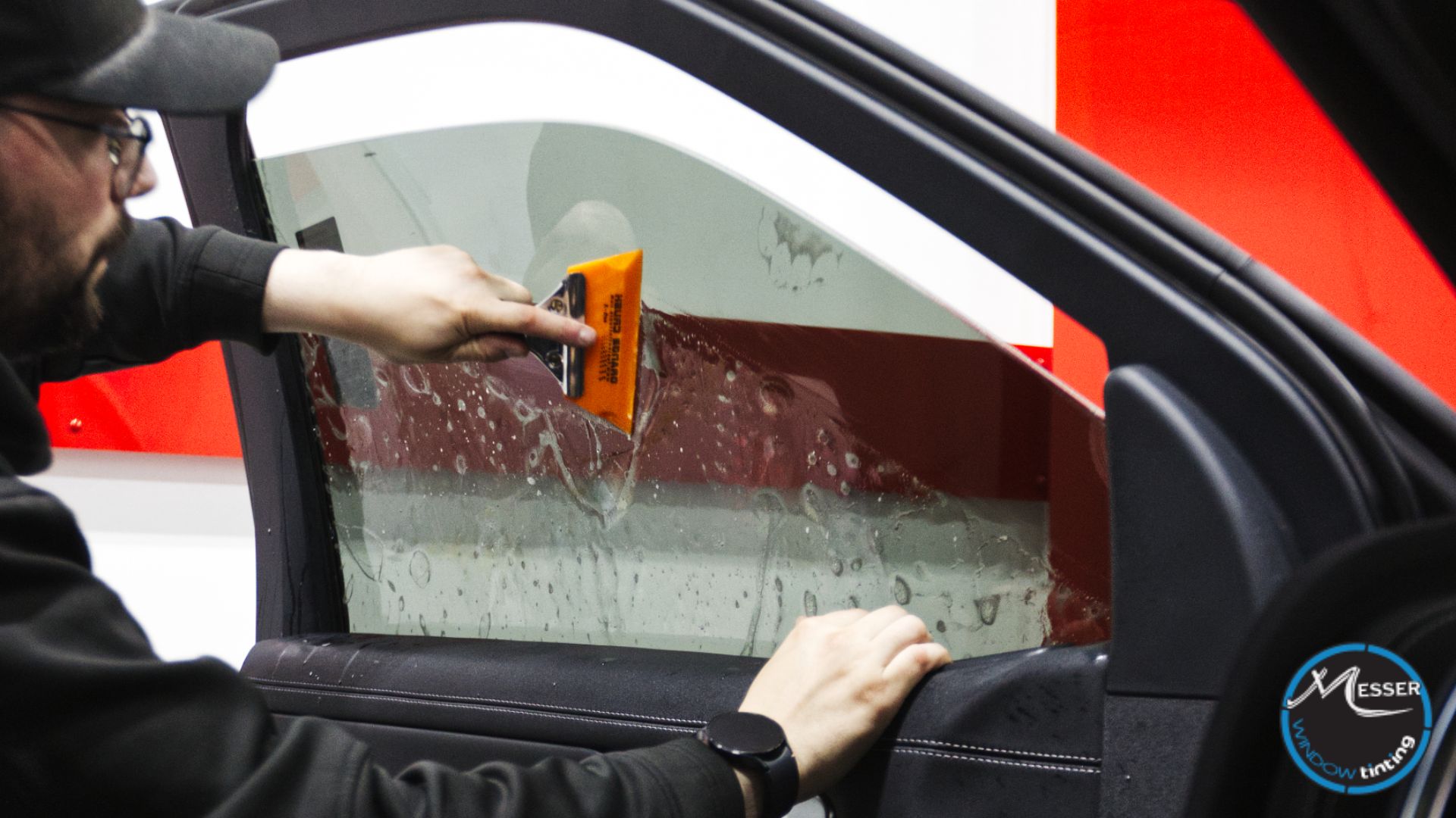 Technician applying window tint film to a car door window using a squeegee to smooth out water and air bubbles during installation. Shop interior visible in the background.