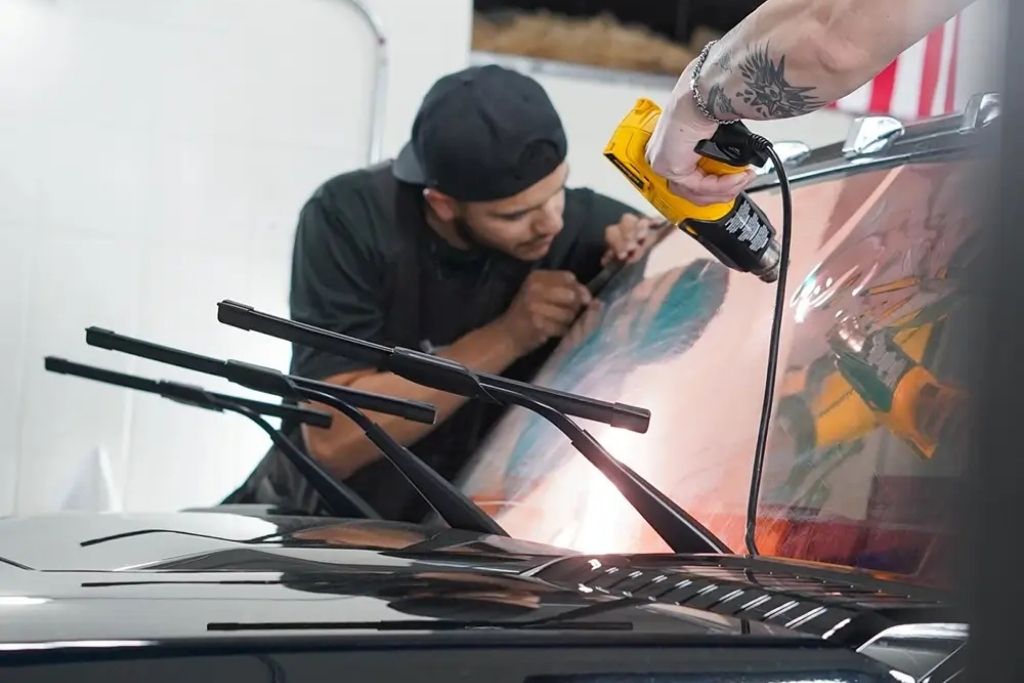 Technician applying automotive window tint to a car windshield using a heat gun during professional installation.