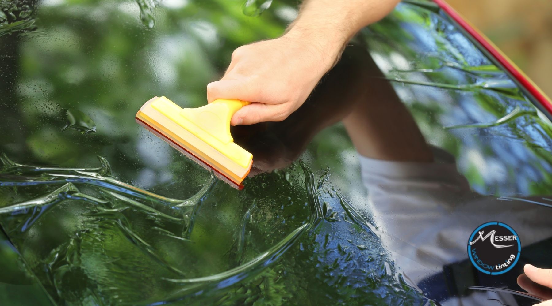 Hand applying window tint film to a car windshield with a yellow squeegee at Messer Window Tinting