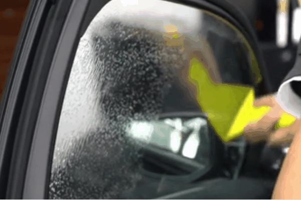 Close-up of a car side window during window tint installation with water droplets and a yellow squeegee smoothing the film